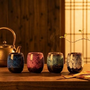 Peut inclure: Quatre tasses à thé en céramique avec un glaçage moucheté dans les tons de bleu, rouge, vert et blanc. Une théière dorée est visible en arrière-plan. Les tasses sont disposées sur une table en bois, suggérant une cérémonie du thé.