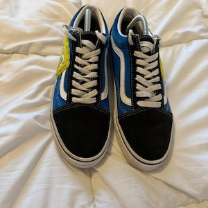 Op de afbeelding: Paar blauwe en zwarte sneakers met witte veters en een witte streep. De sneakers hebben een gele patch aan de zijkant. De schoenen liggen op een wit oppervlak.