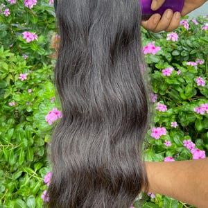 Puede incluir: Un paquete de extensiones de cabello largo, ondulado y de color marrón oscuro con un borde superior tejido. El cabello tiene una textura natural y se sujeta con un peine de plástico morado, mostrando la longitud y el volumen sobre un follaje verde.