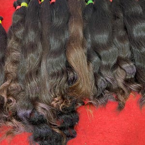 Puede incluir: Una colección de mechones de cabello largo y ondulado en tonos negro y marrón oscuro. Cada mechón está asegurado en la parte superior con una pequeña banda elástica de color rojo, amarillo o verde, manteniendo las hebras alineadas.