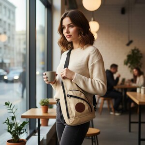 Könnte beinhalten: Beige Sling-Tasche mit schwarzem Riemen und Reißverschluss, mit einem kreisförmigen braunen Aufnäher. Die Tasche wird über der Schulter getragen. Die Person hält einen grauen Becher. Der Hintergrund ist ein Café.