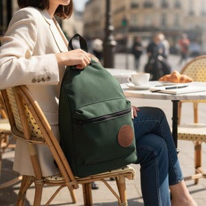 Könnte beinhalten: Ein dunkelgrüner Rucksack mit schwarzem Reißverschluss und einem braunen Lederaufnäher. Der Rucksack hat einen oberen Griff und eine Vordertasche. Die Person sitzt in einem Café mit einer Tasse Kaffee und einem Croissant auf dem Tisch.
