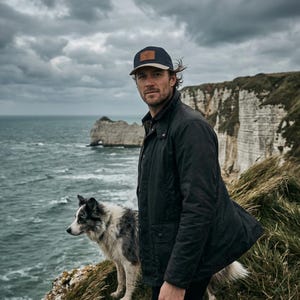 Könnte beinhalten: Eine Person trägt eine marineblaue Baseballkappe mit einem braunen Lederaufnäher, eine dunkle Jacke und eine schwarze Hose und steht mit einem Hund auf einer Klippe. Der Hund ist ein Border Collie mit schwarz-weißem Fell. Der Hintergrund zeigt das Meer und einen bewölkten Himmel.