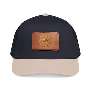 Könnte beinhalten: Marineblaue Baseballkappe mit beigem Schirm und einem braunen Lederaufnäher. Der Aufnäher zeigt eine Illustration eines Jagdhundes und den Text "THE RUSTIC HOUND". Die Kappe hat einen gebogenen Schirm und einen Knopf oben.