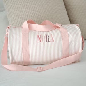 May include: A pink and white striped duffel bag with the name "NORA" in pink lettering. The bag has pink handles and a shoulder strap. The bag is cylindrical and rests on a white surface.