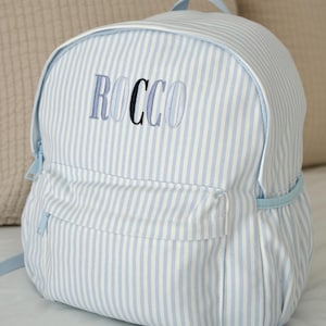 May include: A blue and white striped backpack with the name "ROCCO" embroidered in blue thread. The backpack features a front pocket, a top handle, and adjustable straps. The bag is made of fabric.
