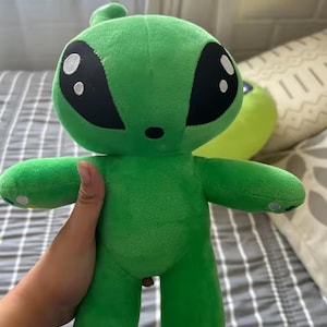 Può includere: Un peluche alieno verde brillante caratterizzato da una grande testa arrotondata con occhi neri a mandorla sovradimensionati contenenti due riflessi circolari bianchi. La morbida creatura imbottita ha un piccolo naso nero, braccia e gambe corte.