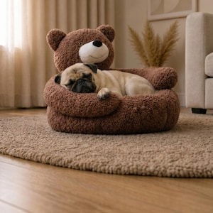 May include: A brown plush pet bed shaped like a teddy bear with a round head, ears, and arms that wrap around the center. A light fawn-colored pug is curled up and sleeping inside the soft, textured fabric of the circular pet bed.