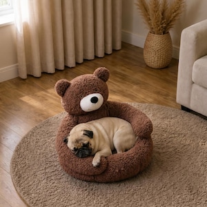 May include: A brown plush pet bed shaped like a teddy bear with a round head, small ears, and a white muzzle. A light fawn-colored pug is curled up inside the soft, textured circular bed, which rests on a matching round brown rug on the floor.