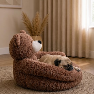 May include: A brown, textured, teddy bear-shaped pet bed with a white muzzle and black nose. A small, tan pug dog is curled up and sleeping inside the soft, plush, rounded structure, which features integrated armrests and a bear head backrest.