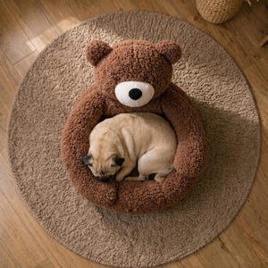 May include: A brown, plush, circular pet bed shaped like a teddy bear with a white muzzle and black nose. The bed features a soft, textured, fleece-like material. A light tan pug is curled up and sleeping inside the center of the bear-shaped bed.