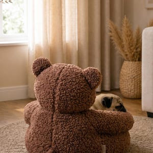 May include: A brown, textured, teddy bear-shaped pet bed with rounded ears and a small tail. The plush, fuzzy material covers the entire exterior of the seat. A small, light-colored pug dog is resting inside the circular, cushioned enclosure.