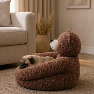 May include: A brown teddy bear shaped pet bed with a textured, plush exterior and a white muzzle. A small fawn colored pug is curled up inside the bed, resting its head on the soft, rounded edge of the bear's body while sleeping peacefully.