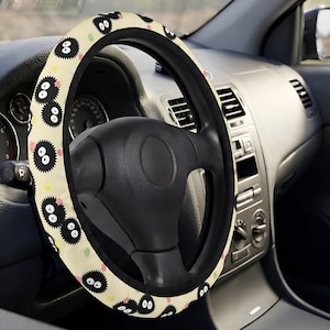 May include: A car steering wheel cover featuring a cream-colored background with a repeating pattern of black, fuzzy, circular soot sprite characters with large white eyes, accented by small pink and yellow star-shaped confetti elements.