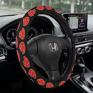 May include: A black steering wheel cover featuring a repeating pattern of red cloud shapes. The cover is fitted onto a black steering wheel with a silver Honda logo in the center, positioned within the interior of a vehicle with a dashboard display.