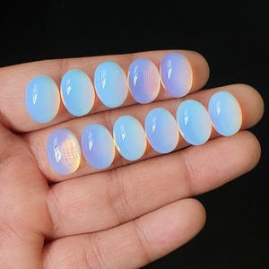 Peut inclure: Dix pierres précieuses cabochons translucides de forme ovale avec une surface lisse et polie. Ces pierres présentent un aspect laiteux bleu pâle avec de subtils reflets irisés qui changent de couleur à la lumière, disposées en deux rangées.