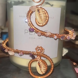Könnte beinhalten: Ein Armreif aus Kupferdraht mit einem Spiraldesign, verziert mit Edelsteinen und einer Muschel. Der Armreif ist um eine Kerze gewickelt, auf deren Etikett die Worte "PEACE HEALING" stehen.