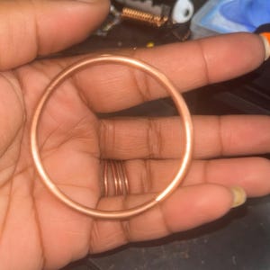 以下が含まれることがあります： 滑らかで光沢のある仕上げの円形銅製バングル。このバングルは、シンプルな連続したバンドデザインが特徴で、円周全体に一貫したメタリックな銅色をしており、すっきりとしたミニマルな美しさを表現しています。