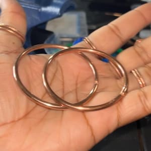 May include: Two circular copper bangles with a smooth, polished finish. The bangles are thin, round, and metallic in appearance, featuring a simple, continuous design. These copper accessories are shown as a pair, highlighting their reflective surface and consistent, uniform thickness throughout the entire circumference of each piece.