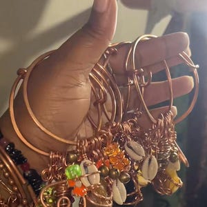 Pode incluir: Uma coleção de pulseiras de fio de cobre com bandas circulares ajustáveis. Cada pulseira é adornada com vários berloques, incluindo pequenas conchas de búzios brancas, contas translúcidas laranja e verdes e esferas metálicas envoltas em fio de cobre.