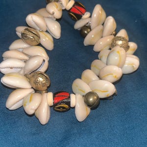 May include: A bracelet constructed from numerous white cowrie shells arranged in a multi-layered pattern, interspersed with metallic gold-toned spherical bells and two cylindrical black beads featuring red and yellow painted geometric patterns.