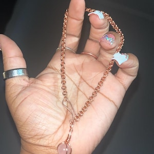 May include: A copper chain necklace featuring a round, light pink gemstone pendant secured in a wire-wrapped setting. The chain is constructed from small, interlocking circular links and includes a simple hook-style clasp mechanism for closure.