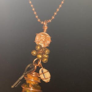 May include: A copper ball chain necklace featuring a pendant assembly with a wrapped penny, a cluster of small brass bells, a metal key, a white cowrie shell, and a miniature glass bottle filled with small items, all secured with copper wire.
