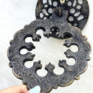 Könnte beinhalten: Verzierte Schubladengriff aus Bronze mit aufwendigen floralen und scrollartigen Details. Der Griff hat eine kreisförmige Rückplatte mit einem dekorativen Muster und einem zentralen Griff. Das Design ist von Vintage inspiriert.