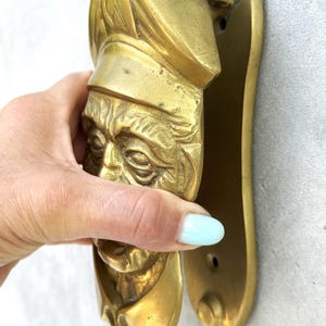 May include: A brass door knocker in the shape of a chef's head, with a detailed face and hat. The knocker is mounted on a matching brass plate. The chef's face is detailed with wrinkles and a stern expression. The knocker is held by a hand.