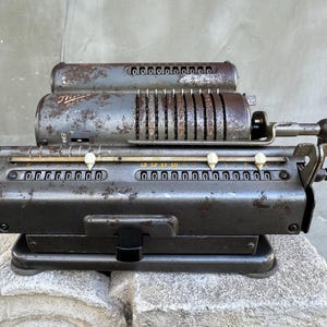 Könnte beinhalten: Ein antiker mechanischer Rechenschieber mit einer verwitterten, grauen Metalloberfläche. Er verfügt über mehrere Reihen von Ziffernrädern, rotierenden Zylindern und einen schwarzen Griff. Der Rechner liegt auf einer Steinoberfläche.