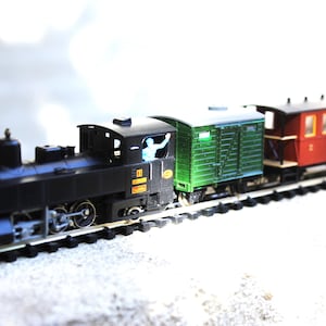 Könnte beinhalten: Ein Modelleisenbahn-Set bestehend aus einer schwarzen Dampflokomotive mit einer kleinen Figur im Führerstand, gefolgt von einem grünen Güterwagen und einem roten Personenwagen mit Fenstern, auf einem schwarzen Kunststoffgleis.