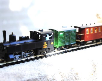 Puede incluir: Un conjunto de tren a escala que consta de una locomotora de vapor negra con una pequeña figura en la cabina, seguido de un vagón de carga verde y un vagón de pasajeros rojo con ventanas, sobre una vía de plástico negra.