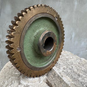 Può includere: Un grande ingranaggio industriale con un anello esterno dentato color bronzo e un disco centrale verde. L'ingranaggio ha un foro centrale ed è montato su una superficie di pietra. I denti dell'ingranaggio sono affilati e ben definiti.