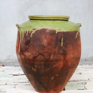 Op de afbeelding: Een grote, ronde voorraadpot van terracotta met een wijde opening en een dikke, lichtgroen geglazuurde rand die uitloopt over het donkere, roodbruine ongeglazuurde lichaam. De pot is voorzien van twee kleine, geïntegreerde handvatten.