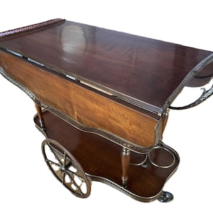 Op de afbeelding: Een houten serveerwagen met een gepolijste donkerbruine afwerking, voorzien van twee uitklapbare zijkanten, een onderste plank en een metalen handvat. De wagen rust op twee grote spaakwielen en twee kleine zwenkwielen met metalen afwerking.