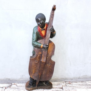 Può includere: Una statuetta dipinta di un musicista che suona un contrabbasso. Il personaggio indossa una giacca verde scuro con colletto rosso e una camicia gialla con papillon. Il contrabbasso è marrone con venature del legno e poggia su una base.