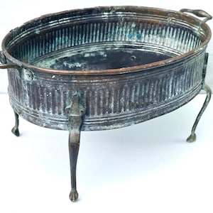 Op de afbeelding: Een ovale koperen bak met een geribbelde buitenkant, voorzien van een verweerde patina met groene oxidatie. De bak heeft twee handvatten aan de zijkanten en rust op vier slanke metalen poten met klauwvormige voeten, met tekenen van ouderdom.