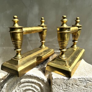 Pode incluir: Dois pares de cães de fogo antigos em latão dourado. Cada cão de fogo tem uma base retangular, uma coluna decorativa e uma barra horizontal. Os cães de fogo são exibidos em uma superfície de pedra. Os cães de fogo são provavelmente usados ​​para segurar toras em uma lareira.