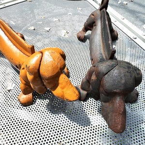 May include: Two cast iron dog sculptures, one orange and one dark gray, rest on a metal surface. The dogs are in a playful pose, with their hindquarters raised and tails curved. The sculptures have a rustic, weathered appearance.