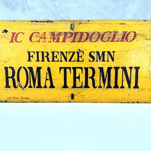 Könnte beinhalten: Ein rechteckiges gelbes Zugschild aus Metall mit schwarzer und roter Schrift. Das Schild trägt die Aufschrift IC CAMPIDOGLIO, FIRENZE SMN und ROMA TERMINI. Es hat eine abgenutzte, zerkratzte Oberfläche mit Befestigungslöchern an den Seiten.