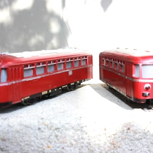 Op de afbeelding: Twee rode modeltreinrijtuigen met een reeks rechthoekige ramen langs de zijkanten, een zilverkleurig dak en zwarte onderstellen. Elk rijtuig heeft een voorruit, kleine ronde koplampen en gedetailleerde markeringen op de zijkant.