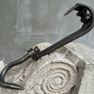 May include: A dark metal hook with a curved design, featuring a pointed end and a pivoting joint. The hook is displayed against a textured, light-colored stone surface, showcasing its industrial aesthetic.