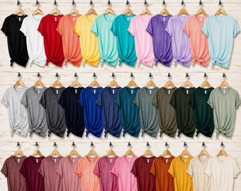 以下が含まれることがあります： 木製のハンガーに掛けられた、様々な無地の半袖クルーネックTシャツ45枚のコレクション。Tシャツは明るい木目調の背景に対して、3つの水平な列に並べられています。