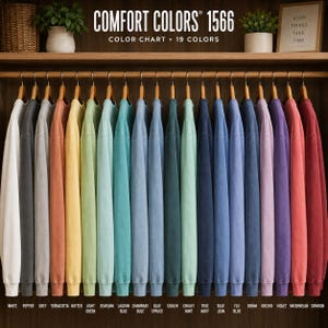 May include: A collection of nineteen long-sleeve sweatshirts from Comfort Colors 1566 displayed on a wooden rack. The garments are arranged in a color gradient ranging from white, grey, and terracotta to various shades of blue, green, purple, and red.