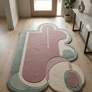 May include: An area rug featuring an irregular, organic shape with a tufted texture. The design consists of layered, rounded geometric sections in muted shades of dusty rose, sage green, and cream, creating a multi-dimensional, abstract pattern.