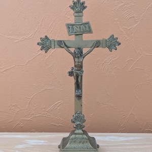 May include: A metallic bronze-toned standing crucifix featuring a central figure of Jesus on the cross. The cross arms and top are adorned with decorative fan-shaped finials. A small plaque above the figure is inscribed with the letters INRI.