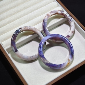 May include: Three circular bangles crafted from polished stone, featuring a marbled pattern of deep purple, soft lavender, creamy white, and translucent grey tones. Each bangle has a smooth, rounded surface with natural variations in color.