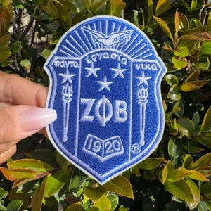 May include: A blue and white embroidered patch with the letters "ZΦB" and the year "1920" in white. The patch features a white bird with outstretched wings, three white stars, and two white torches.