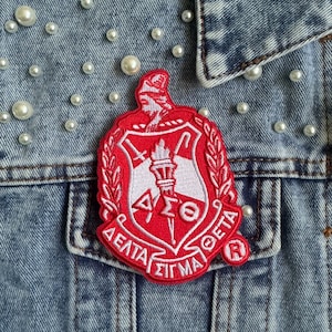 May include: A red and white embroidered patch with the Greek letters Delta Sigma Theta and a white shield with a torch and other symbols. The patch is on a blue denim jacket with white pearl-like embellishments.