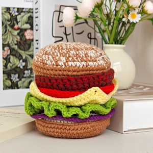 May include: A crocheted hamburger toy featuring a tan bun, a dark brown patty, a bright red tomato slice, a yellow cheese slice, ruffled green lettuce, and a purple onion slice, all stacked together in a realistic layered food construction.
